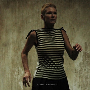 May include: A woman wearing a black and white striped sleeveless shirt with a black bow on each shoulder. The shirt has a boat neck and is tucked into black pants. The woman is standing in front of a textured wall. The text "PEOPLE'S COUTURE" is printed at the bottom of the image.