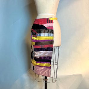 Puede incluir: Una falda hasta la rodilla confeccionada con paneles horizontales de satén y tela texturizada multicolor, en tonos rosa, amarillo, morado, marrón y negro. La cintura se asegura con una correa de color amarillo brillante y herrajes metálicos.