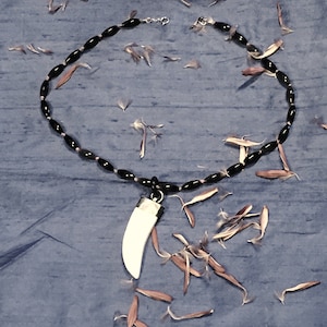 May include: A black beaded necklace with a white tooth pendant. The necklace is lying on a blue surface with white flower petals scattered around it.