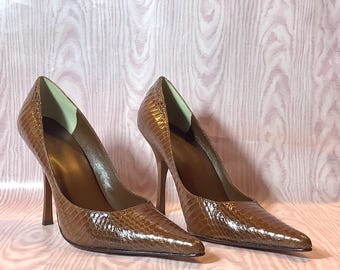 May include: A pair of brown high-heeled shoes with pointed toes. The shoes have a textured, reptile-like pattern and slender heels. The interior lining is a lighter shade of brown. The shoes are displayed on a pink surface.