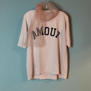 Może przedstawiać: Jasnoróżowy T-shirt z krótkim rękawem z napisem AMOUR wydrukowanym pogrubionymi czarnymi wielkimi literami na klatce piersiowej. Przezroczysty, bladoróżowy szal z organzy jest udrapowany wokół dekoltu koszulki, która wisi na wieszaku.