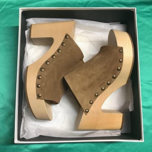May include: A pair of light brown wooden platform clogs with a suede upper in a box. The clogs have a peep-toe design and are adorned with small, round, gold-colored studs along the edges. The box is white with a black interior.