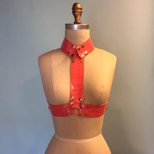 Puede incluir: Un top halter de cuero rosa con tachuelas doradas. El top tiene un escote alto y un diseño de tirantes en forma de Y.