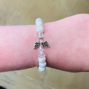 May include: White beaded bracelet with a silver angel charm. The beads are iridescent and have a subtle shimmer.
