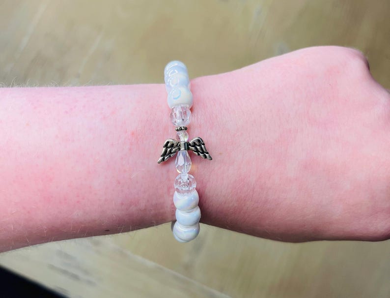 May include: White beaded bracelet with a silver angel charm. The beads are iridescent and have a subtle shimmer.