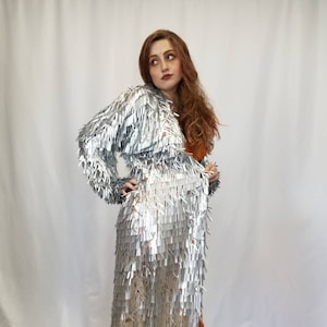 May include: A silver fringe jacket and matching long skirt. The outfit is paired with platform shoes with a floral design and a bright orange accent. The outfit is a unique, eye-catching ensemble.