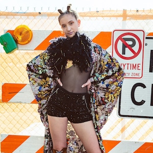 May include: A woman in a sequined kimono, black shorts, and brown lace-up boots. The kimono has a multi-coloured floral pattern. The boots are knee-high. The background features a construction zone with orange and white barriers.