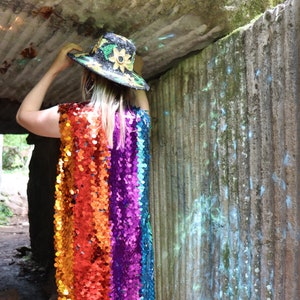 May include: A long, sleeveless sequin dress in a rainbow of colors, from red to blue. The dress is paired with white boots and a black hat decorated with a yellow sunflower and green leaves. The outfit is eye-catching and vibrant.