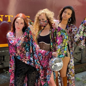 May include: Three people wearing colourful sequined jackets. The person in the middle is wearing a black tank top and a face mask. The person on the right is holding a silver disco ball.