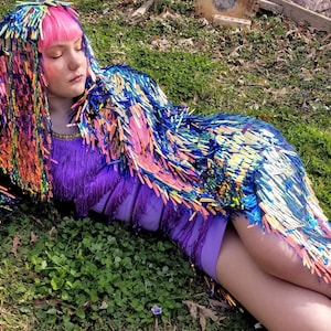 May include: A woman wearing a colorful sequined jacket with a hood, a purple dress, and silver boots. She is lying on her side in a grassy field.