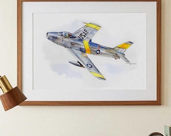 May include: A framed watercolor painting of a silver jet aircraft with yellow wing and fuselage accents. The plane features the text USAF on the side and a star insignia on the wing. The artwork is displayed in a brown wooden frame on a wall.