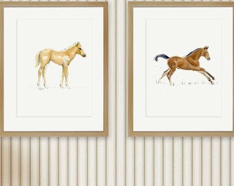 May include: Two framed wall art prints featuring watercolor illustrations of horses. The left print shows a light tan foal standing, while the right print depicts a brown horse galloping. Both artworks are set in light wood frames with white mats.