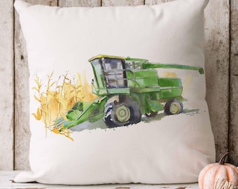 May include: A square white throw pillow featuring a watercolor illustration of a green combine harvester working in a field of golden yellow corn. The artwork displays the agricultural machinery in profile with visible wheels and a header.