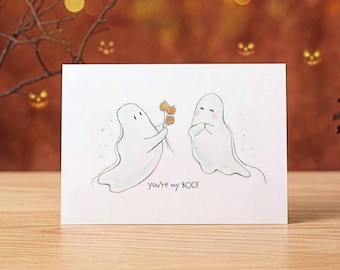 May include: A white greeting card featuring a hand-drawn illustration of two ghosts. One ghost offers three small orange pumpkins to the other. The text "you're my BOO!" is written in black ink below the ghosts on the white card surface.