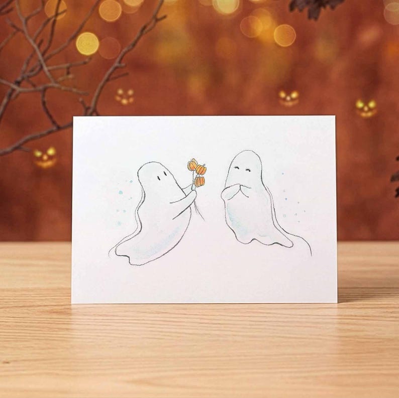May include: A white rectangular greeting card featuring a hand-drawn illustration of two white ghosts. One ghost on the left holds a small bouquet of three orange pumpkins, while the other ghost on the right stands with hands clasped together.