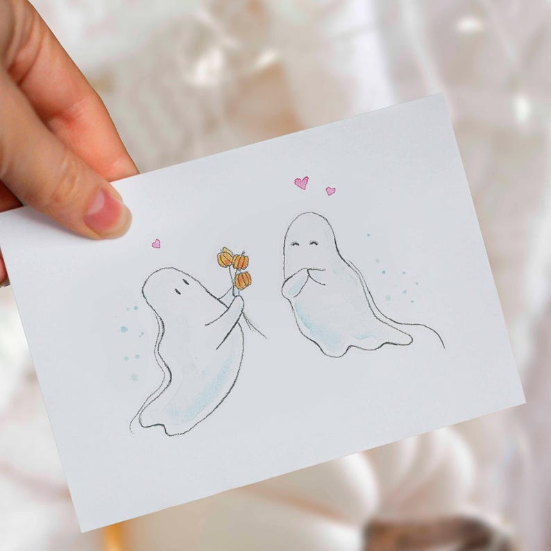 May include: A white greeting card featuring a hand-drawn illustration of two ghosts. One ghost offers three small orange pumpkins to the other ghost. Three small pink hearts are drawn above the ghosts, and light blue dots surround them.