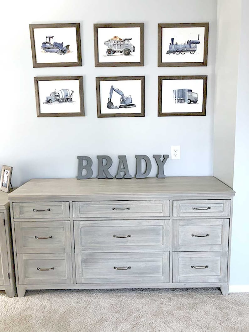 customer lifestyle photo showing blue bulldozer, gray dump truck, navy blue train, blue gray cement mixer truck, blue gray excavator and blue gray garbage truck hung in a toddler boys room.