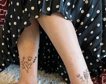 May include: Two black ink floral temporary tattoos applied to the lower legs. Each tattoo features a delicate botanical illustration of wildflowers and stems with fine lines. The tattoos are positioned on the skin above the ankles.