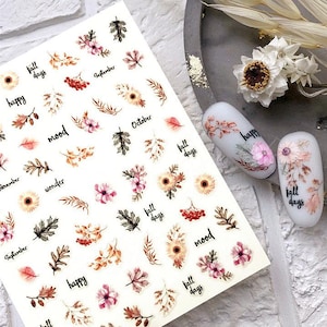 Peut inclure: Autocollants pour nail art avec des motifs d'automne. La feuille présente des illustrations florales et de feuilles en marron, rose et orange, avec des textes comme "happy", "mood" et "fall days". Deux faux ongles décorés sont également visibles.