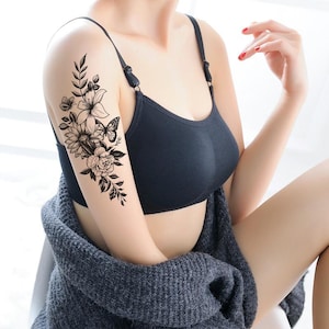May include: A black and white floral temporary tattoo on an arm, including a sunflower, roses, and a butterfly. The person is wearing a black bra and a grey sweater. The tattoo design is detailed.