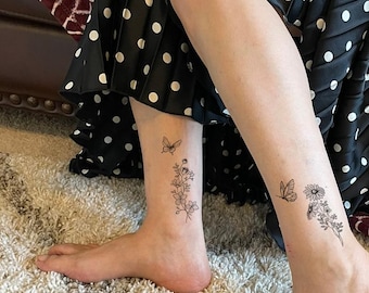 May include: Two temporary tattoos applied to the lower legs, each featuring a fine-line black ink illustration of a small butterfly hovering above a cluster of wildflowers, including daisies and leafy stems, rendered in a delicate botanical style.