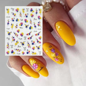 Puede incluir: Hoja de pegatinas de arte de uñas con diseños florales coloridos en amarillo, rosa y azul. La hoja incluye texto pequeño con palabras como "Wander" y "Simple". La imagen también muestra uñas pintadas de amarillo con diseños florales.