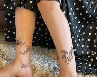 May include: Black ink temporary tattoos applied to the lower legs. One leg features a butterfly, a crescent moon, and a small wave design. The other leg displays a cluster of wildflowers with stems and leaves. The tattoos are fine-line black ink.