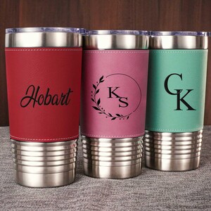 Puede incluir: Tres vasos de acero inoxidable con diferentes envolturas de cuero. Los vasos est&aacute;n personalizados con los nombres "Hobart", "KS" y "CK".