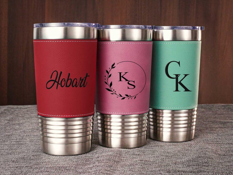 Puede incluir: Tres vasos de acero inoxidable con diferentes envolturas de cuero. Los vasos est&aacute;n personalizados con los nombres "Hobart", "KS" y "CK".