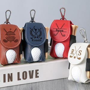 May include: Leather golf ball holders in brown, navy blue, red, and white, each with a golf ball and tees. The holders have personalized text and golf club designs, attached to a silver keychain.