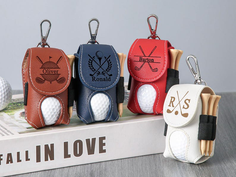 May include: Leather golf ball holders in brown, navy blue, red, and white, each with a golf ball and tees. The holders have personalized text and golf club designs, attached to a silver keychain.