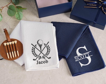 May include: Golf-themed gift set featuring a white waffle-weave towel with a golf ball and club design, and the name "Jacob". A navy blue towel with the name "Scott" is also present, along with a leather golf tee holder and a gift box.