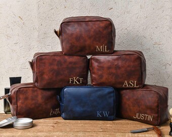 Puede incluir: Un conjunto de neceseres de cuero marrón con iniciales bordadas en dorado. Una bolsa azul está en el centro. Accesorios de aseo, como una brocha de afeitar y una navaja, están sobre una superficie de madera.