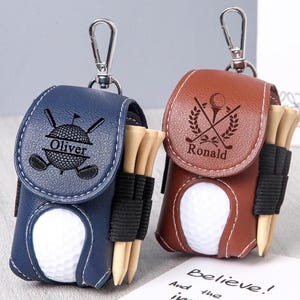 May include: Two personalized golf ball holders. One is navy blue with "Oliver" engraved, the other is brown with "Ronald". Each has a golf ball, tees, and a silver clip.