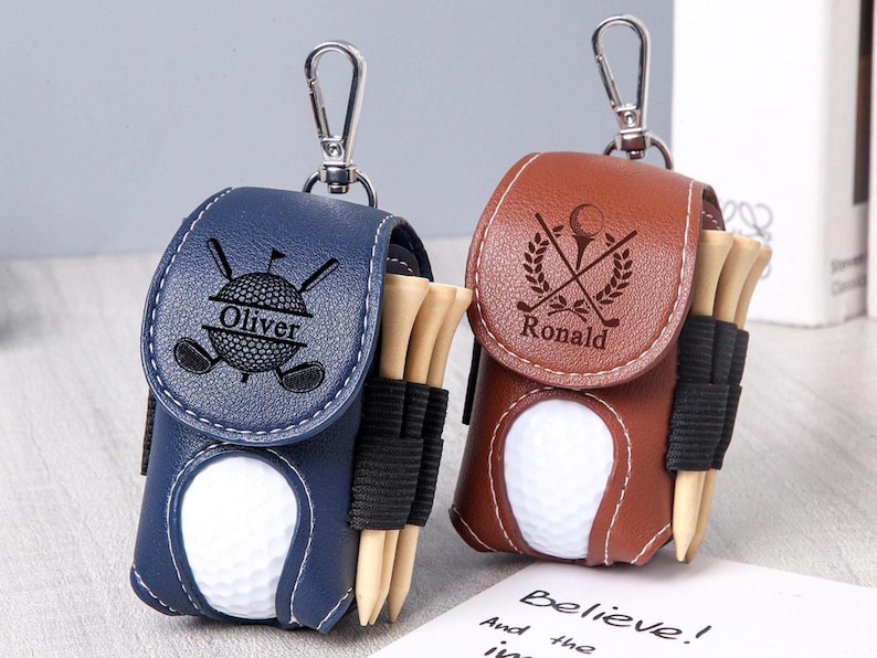 May include: Two personalized golf ball holders. One is navy blue with "Oliver" engraved, the other is brown with "Ronald". Each has a golf ball, tees, and a silver clip.