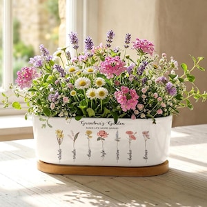 May include: A white ceramic planter filled with a variety of colorful flowers, including pink, purple, and white blooms. The planter has the text "Grandma's Garden" and floral illustrations. It sits on a wooden base, placed on a wooden table.