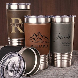 Puede incluir: Tres vasos de acero inoxidable con envolturas de cuero. Los vasos están personalizados con los nombres "Robert", "Richard" y "Jacob". El vaso con el nombre "Jacob" también tiene el texto "Groomsman 25.04.2025".