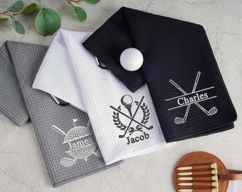 May include: Golf towels in gray, white, and black with personalized embroidery. Each towel features a golf-themed design and a name. A golf ball rests on one of the towels. A leather golf tee holder is also visible.
