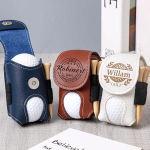 May include: Three leather golf ball holders in navy blue, brown, and white. Each holder contains a golf ball and wooden tees. The navy blue holder is open, while the others are closed. The holders have personalized text.