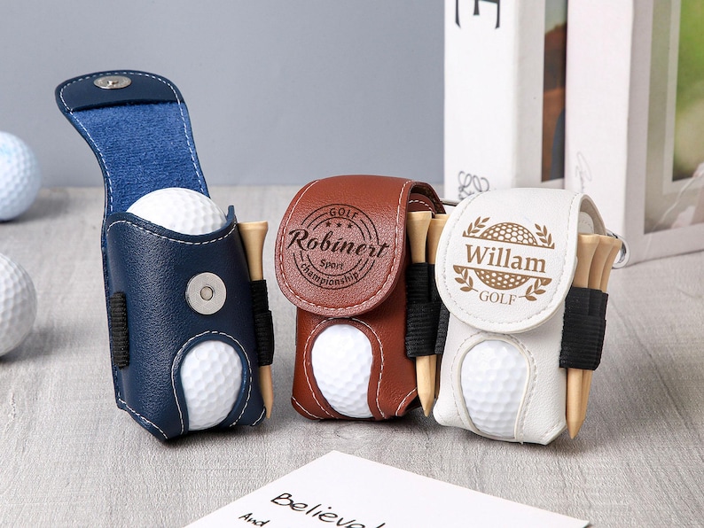 May include: Three leather golf ball holders in navy blue, brown, and white. Each holder contains a golf ball and wooden tees. The navy blue holder is open, while the others are closed. The holders have personalized text.