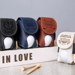 May include: A collection of personalized golf ball holders in black, navy blue, brown, and white leather. Each holder features a golf ball and tee, with names like "Ronald," "Barton," "Clement," and "Willam" engraved on them. The holders are displayed on a white surface.
