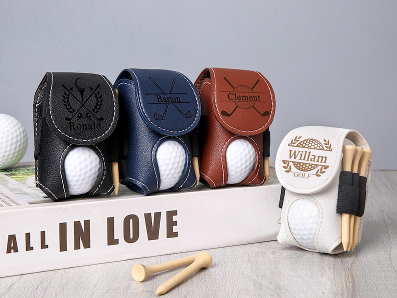 May include: A collection of personalized golf ball holders in black, navy blue, brown, and white leather. Each holder features a golf ball and tee, with names like "Ronald," "Barton," "Clement," and "Willam" engraved on them. The holders are displayed on a white surface.