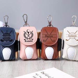 May include: Four personalized golf ball holders in navy blue, pink, brown, and white leather. Each holder has a silver clip, a golf ball, and wooden tees. The holders are engraved with names and golf-themed graphics.