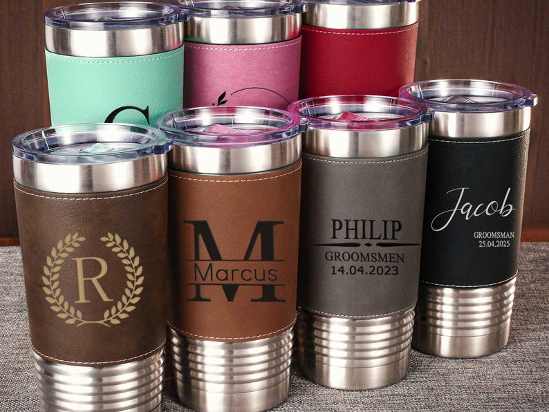 Puede incluir: Conjunto de ocho vasos de acero inoxidable personalizados con envolturas de imitaci&oacute;n de cuero. Cada vaso presenta un nombre y un dise&ntilde;o diferentes, incluidas iniciales, una corona y las palabras "Groomsmen" y "Jacob".