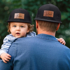 May include: Two black baseball caps with brown leather patches. One cap reads "Kaia" and the other reads "Dad". The caps are worn by a child and an adult. The child is being held by the adult.