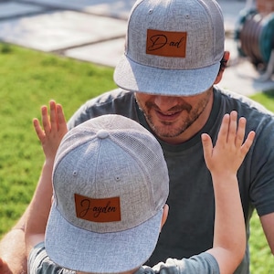 Könnte beinhalten: Graue Baseballkappen mit Mesh-Rücken, hellgrauem Schirm und einem braunen Lederpatch. Eine Kappe hat das Wort "Dad" und die andere den Namen "Jayden" in Schreibschrift. Die Kappen werden von einem Vater und seinem Kind getragen.