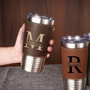 Puede incluir: Tres vasos de acero inoxidable personalizados con envolturas de cuero. Los vasos son marrones, grises y marrones. Los nombres "Marcus", "Robert" y "Baron" est&aacute;n grabados en los vasos.