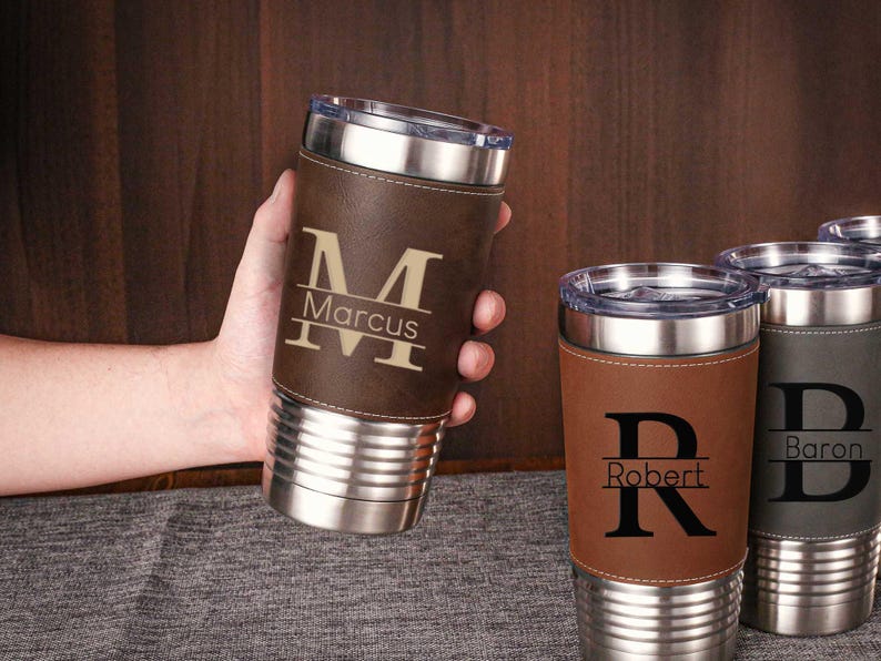 Puede incluir: Tres vasos de acero inoxidable personalizados con envolturas de cuero. Los vasos son marrones, grises y marrones. Los nombres "Marcus", "Robert" y "Baron" est&aacute;n grabados en los vasos.