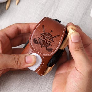 May include: A brown leather golf ball holder with a golf ball and tees. The holder has a golf-themed design with the name "Oliver" and a golf ball, clubs, and flag. The holder is held in a person's hands.