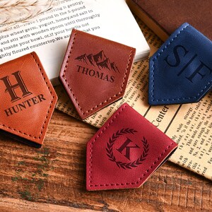 May include: Four leather bookmarks in shades of brown, red, and blue, each with a unique engraved design. One bookmark features the name "HUNTER", another "THOMAS", and a third with the initials "S/F". The fourth has a "K" with a laurel wreath.